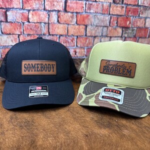 May include: Two trucker hats with leather patches. One is black with "SOMEBODY" on the patch. The other is olive green and camouflage with "Somebody's PROBLEM" on the patch. Both have mesh backs and adjustable straps.