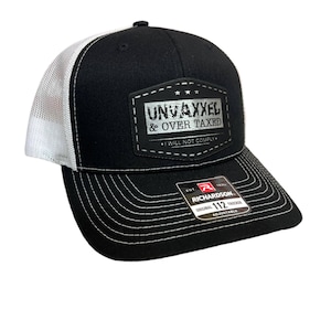 May include: Black and white trucker hat with a white mesh back. The hat has a black patch with the text "UNVAXXED & OVER TAXED I WILL NOT COMPLY" in silver lettering. The hat is a Richardson 112 Original Trucker.