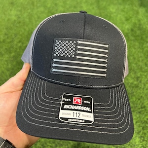 May include: Black and gray trucker hat with a white American flag design featuring wrenches. The hat has a black mesh back and a gray brim. The hat has a red, white, and black label that reads "EST. 1970 RICHARDSON. ORIGINAL 112 TRUCKER ADJUSTABLE".