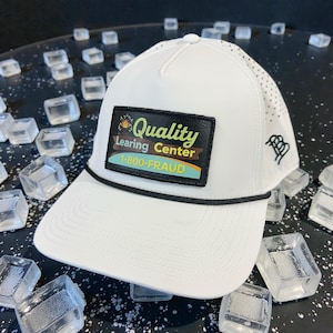 May include: White baseball cap with a black rope detail and a rectangular patch. The patch features the words "Quality Learing Center" and "1-800-FRAUD". The cap has a curved brim and a small logo on the side.