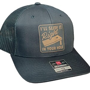 May include: A dark gray trucker hat with a brown leather patch that says "I'll slide it right in your hole" with a cornhole board graphic. The hat is adjustable and has a mesh back.