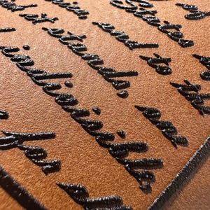 May include: Close-up of a brown leather surface with raised black lettering. The text appears to be a quote or passage from a book. The leather has a textured appearance, and the lettering is slightly glossy.