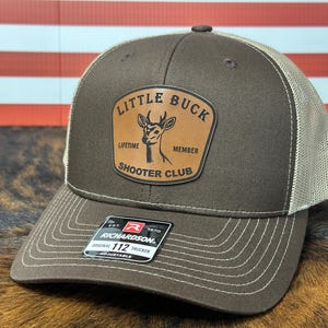 May include: A brown and tan trucker hat with the text "Little Buck Shooter Club" on a leather patch. The hat has a mesh back and a curved brim. The Richardson 112 adjustable hat is displayed on a brown surface.