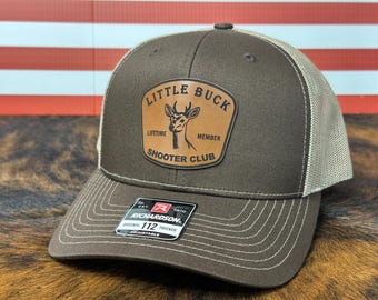 May include: A brown and tan trucker hat with the text "Little Buck Shooter Club" on a leather patch. The hat has a mesh back and a curved brim. The Richardson 112 adjustable hat is displayed on a brown surface.