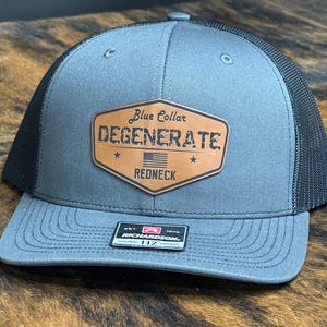May include: A gray and black trucker hat with a brown leather patch that says "Blue Collar Degenerate Redneck" with a small American flag in the center.