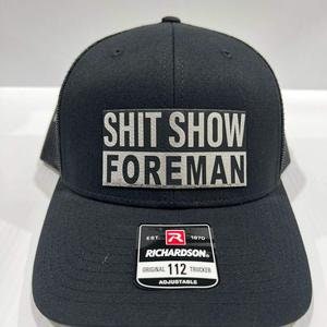 May include: A black and grey trucker hat with a white patch that reads "SHIT SHOW FOREMAN". The hat is adjustable and has a Richardson logo.