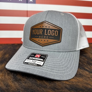 May include: Gray trucker hat with a white mesh back and a brown leather patch. The patch features the text "YOUR LOGO HERE" and stars. The hat has a curved brim and an adjustable strap.