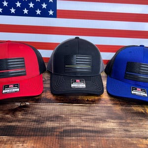 May include: Three trucker hats with black mesh back panels. The hats are red, black, and blue. Each hat features a black American flag with a thin colored stripe across it. The red hat has a red stripe, the black hat has a green stripe, and the blue hat has a blue stripe.