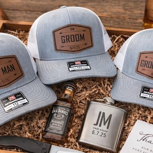 May include: Three light gray and white mesh trucker hats with brown leather patches featuring the text Best Man, Groom, and Groomsman, alongside a metal flask engraved with JM 6.7.25, a miniature bottle of whiskey, a pocket knife, and a cigar.