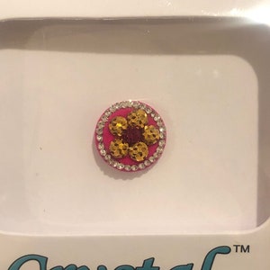 May include: A pink and gold flower-shaped bindi with rhinestones around the edge. The bindi is on a white background with the text "Crystal Designer" below it.