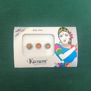 May include: A package of three decorative bindis in gold, red, and gold with white accents. The package has the text "RIM ZIM" and "Kusum FANCY BINDI" with an illustration of a woman in traditional Indian attire.