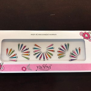 May include: A white box with the text "sign of successful women" and the brand name "yaSSvi". The box contains colorful, fan-shaped face jewels with rhinestones. The jewels are arranged in a decorative pattern.