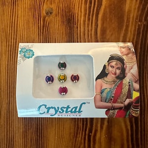 May include: A pack of six colorful bindi stickers with rhinestones. The bindi stickers are arranged in a circle on a white background. The pack is labeled "Crystal Designer".