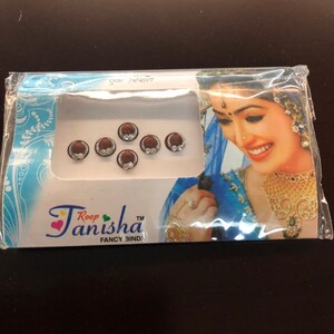 May include: A package of Roop Tanisha Fancy Bindi. The package contains six round, dark red bindis with a silver-colored border and a clear, sparkling center. The packaging features a woman in traditional Indian attire.