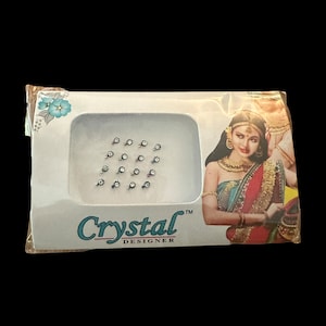 May include: A white package with a clear plastic window showing a sheet of 12 small, clear rhinestones with silver settings. The package has the text "Crystal Designer" printed on it.