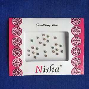May include: A white and pink card with the text "Something New" and "Nisha™" on it. The card contains a sheet of small, colorful, iridescent rhinestones.