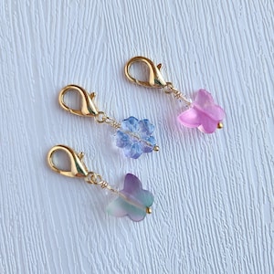 May include: Three gold-colored lobster clasp charms with delicate floral and butterfly pendants. The pendants are in shades of blue, purple, and pink, with clear bead accents. The charms are on a textured white surface.