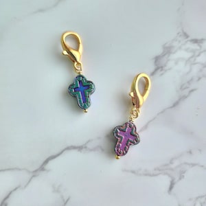 May include: Two gold-colored charm clasps each holding a cross-shaped charm. The crosses are iridescent with shades of blue, green, and purple. Each charm has a small gold bead at the bottom.