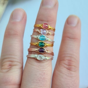 May include: A stack of five wire-wrapped rings with different colored gemstones. The rings are stacked on a finger, with the bottom ring featuring a clear gemstone, followed by a purple gemstone, a green gemstone, a blue gemstone, and a pink gemstone.