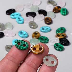 Puede incluir: Una colección de botones ovalados en varios colores y materiales. Los botones incluyen turquesa, malaquita, ojo de tigre y piedra transparente. Algunos botones tienen dos agujeros para coser. Los botones están dispersos sobre una superficie gris claro.