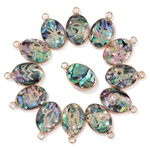 Könnte beinhalten: Ovale Anhänger aus Abalone-Muscheln mit goldfarbenen Rändern, die jeweils ein anderes Tierkreiszeichen zeigen. Die schillernden Muscheln zeigen eine Reihe von Farben, darunter Grün, Blau und Lila. Diese Anhänger sind für die Schmuckherstellung bestimmt.