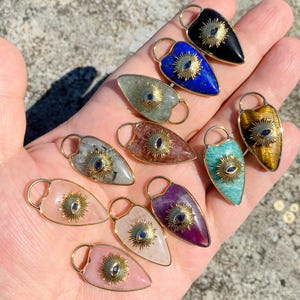 Könnte beinhalten: Eine Sammlung von Edelsteinanhängern in verschiedenen Farben, darunter Rosa, Lila, Blau und Grün. Jeder Anhänger hat ein goldfarbenes Sonnenstrahlendesign mit einem blauen Auge in der Mitte. Die Anhänger sind tropfenförmig und haben eine kleine Schlaufe oben.