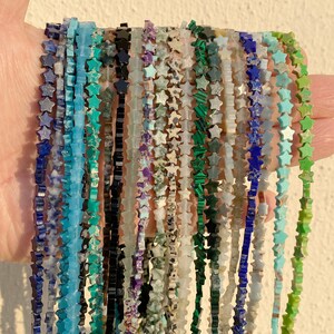 Puede incluir: Una colección de collares de cuentas, cada uno con cuentas en forma de estrella en varios colores. Los collares se sostienen en una mano, mostrando los diversos tonos de las piedras preciosas, incluyendo azul, verde, negro y blanco. Las cuentas miden aproximadamente 1,3 cm.