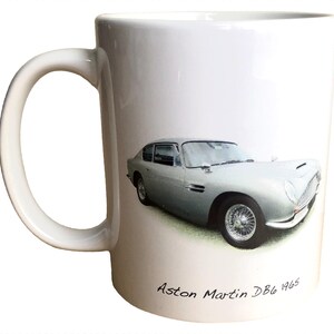以下が含まれることがあります： 1965年製のシルバーのアストンマーティンDB6が描かれた白いセラミックマグカップ。車は詳細なイラストで表現されています。マグカップには持ちやすいように湾曲したハンドルが付いています。
