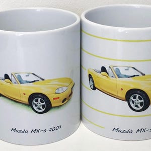 May include: Two white ceramic mugs featuring a yellow Mazda MX-5 convertible car design. One mug has the text "Mazda MX-5 2003" and the other has "Mazda MX-5". The car is yellow with black wheels.
