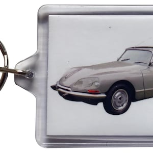 Puede incluir: Un llavero de acrílico transparente con un coche clásico gris claro. El coche tiene un diseño elegante con un techo oscuro y está sobre un fondo blanco. El llavero incluye un anillo de metal plateado para sujetar las llaves.