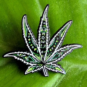 May include: A decorative pin in the shape of a cannabis leaf. The pin features a black outline with intricate geometric patterns in shades of green and white. The pin is set against a green background.