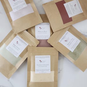 Op de afbeelding: Zes bruine papieren zakjes met verschillende soorten kleimaskers voor het gezicht. De zakjes zijn gelabeld met de namen van de maskers: Super Berry, Mega Greens, Hibiscus Rose, Java Cacao, Matcha Oats en Turmeric Calendula.