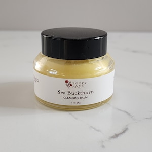 May include: A small glass jar with a black lid contains a yellow balm. A white label wraps around the jar, displaying the text "Sea Buckthorn Cleansing Balm" and the Poppy Lane Organics logo. The jar is 2 oz / 56g.