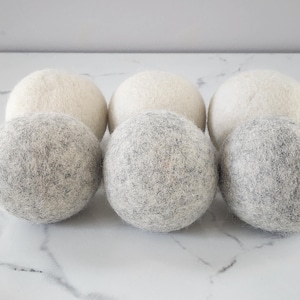 May include: Six wool dryer balls, three are white and three are gray. The balls are arranged in a row on a white marble surface.