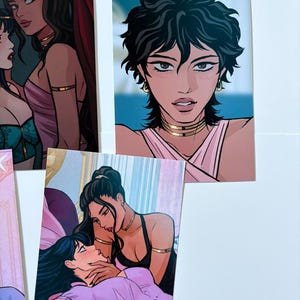 以下が含まれることがあります： 女性のイラストが描かれたアートプリントがいくつかあります。プリントには、金色のジュエリーとピンクまたは黒の服を着た、黒髪の女性が描かれています。アートスタイルは漫画のイラストを彷彿とさせます。