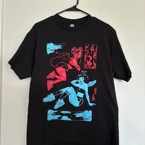 May include: Black t-shirt featuring a red and blue graphic of a devil and an angel. The devil is depicted with horns and a tail, while the angel has wings. Both figures are on clouds, with flames and cloud details.
