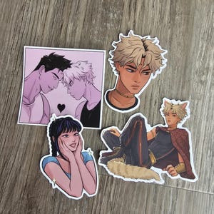 Può includere: Quattro adesivi con illustrazioni in stile anime. Uno mostra due figure con un cuore, un altro una testa maschile e altri due figure in varie pose. Gli adesivi hanno bordi bianchi e sono su una superficie di legno.