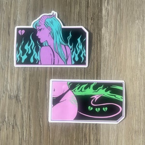 May include: Two stickers with a devil theme. One features a purple demon with teal hair and horns against a black background with teal flames. The other shows a pink figure with a black bikini, green flames, and broken heart graphics.