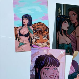 以下が含まれることがあります： 黒髪の女性のイラストが描かれた複数のアートプリント。1つのプリントは虎と女性、別のプリントは2人の女性、3つ目は笑顔の女性が描かれています。プリントは漫画のスタイルです。