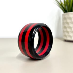 May include: A wide, circular bangle bracelet featuring a high-gloss finish with alternating vertical stripes in solid black and vibrant red. The bracelet has a smooth, rounded exterior surface and a hollow centre, showcasing a bold, two-tone colour pattern.