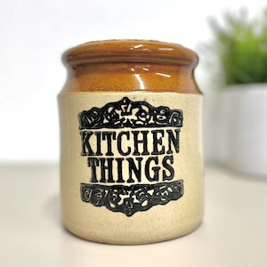 May include: A ceramic kitchen storage jar with a brown lid and a beige body. The jar has the words "KITCHEN THINGS" in black, decorative lettering. A small white pot with a green plant is in the background.