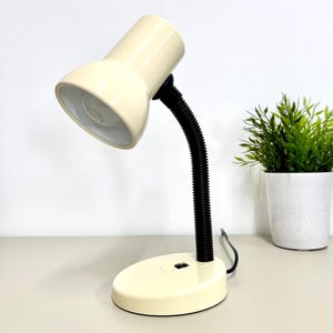 Puede incluir: Lámpara de escritorio color crema con cuello y base negros y flexibles. La lámpara tiene una pantalla redondeada y un pequeño interruptor de encendido/apagado. Una pequeña planta en maceta está a la derecha de la lámpara. La lámpara está sobre una superficie clara.
