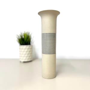 Può includere: Un vaso cilindrico alto in ceramica con una finitura opaca color bianco sporco. Il vaso presenta un bordo svasato nella parte superiore e una fascia centrale di scanalature orizzontali grigie incise che avvolgono la sezione centrale del corpo.