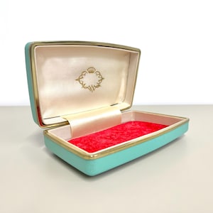 Puede incluir: Caja de joyería rectangular abierta, turquesa claro con ribete dorado. El interior presenta una base de terciopelo rojo y una tapa crema con un emblema dorado. Diseñada para guardar objetos pequeños.