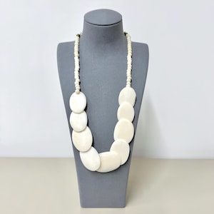 Puede incluir: Un collar que presenta una hilera de pequeñas cuentas redondas de color blanco roto que pasan a una serie de discos ovalados más grandes, lisos y de color blanco roto, dispuestos en cascada. El collar se muestra sobre un busto gris.