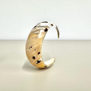 Puede incluir: Un brazalete curvo y abierto con un patrón moteado. El brazalete tiene un color base marrón claro con manchas negras y blancas. La forma general es una forma de C suave y redondeada.