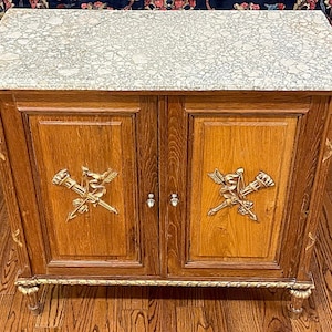 May include: A wooden cabinet with two doors and a marble top. The doors are decorated with gold accents and floral motifs. The cabinet is standing on a wooden floor.
