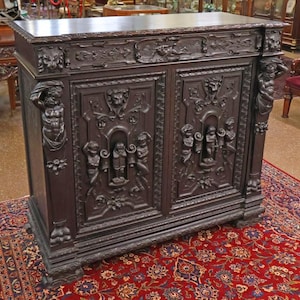 May include: Dark brown antique cabinet with intricate carvings. The cabinet features detailed relief sculptures of figures and floral designs on the doors and sides. The cabinet is set on a patterned red rug.