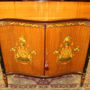 May include: A wooden cabinet with two doors, painted with floral designs and gold accents. The cabinet has a curved top and sits on four legs.