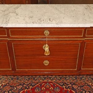 May include: A mahogany-colored cabinet with a white marble top. The cabinet has gold trim and hardware, including drawer pulls and decorative accents. The piece has a classic, elegant design.